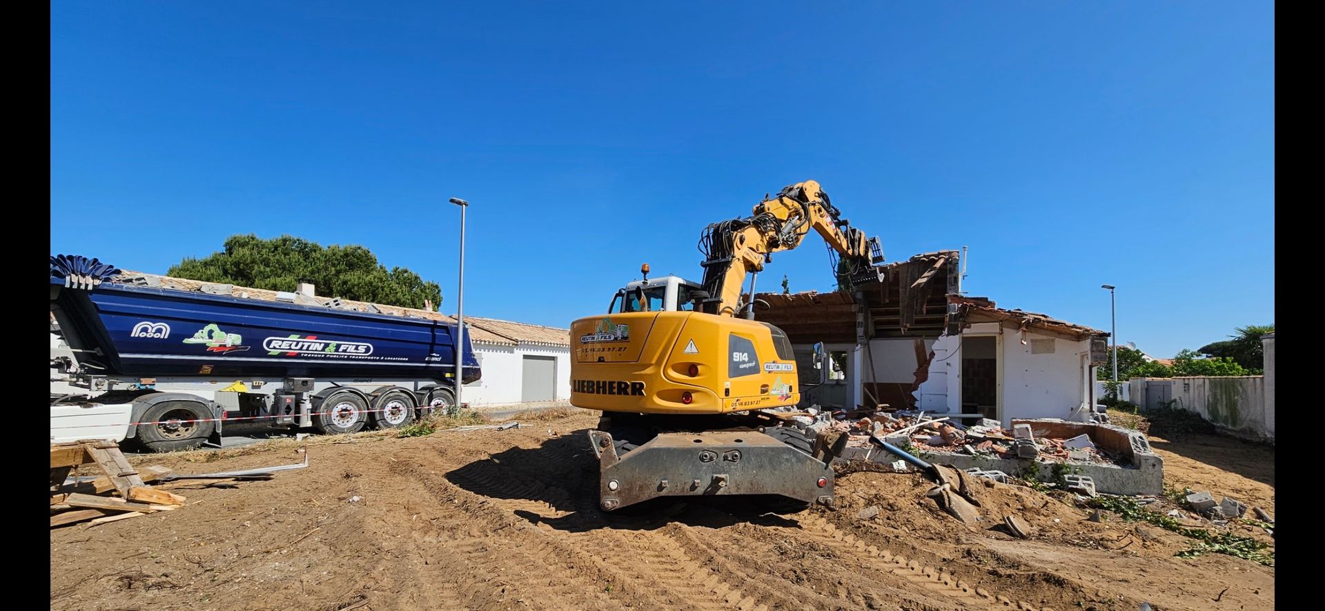 Une pelleteuse démolit un bâtiment ; un camion se trouve à proximité par une journée ensoleillée.