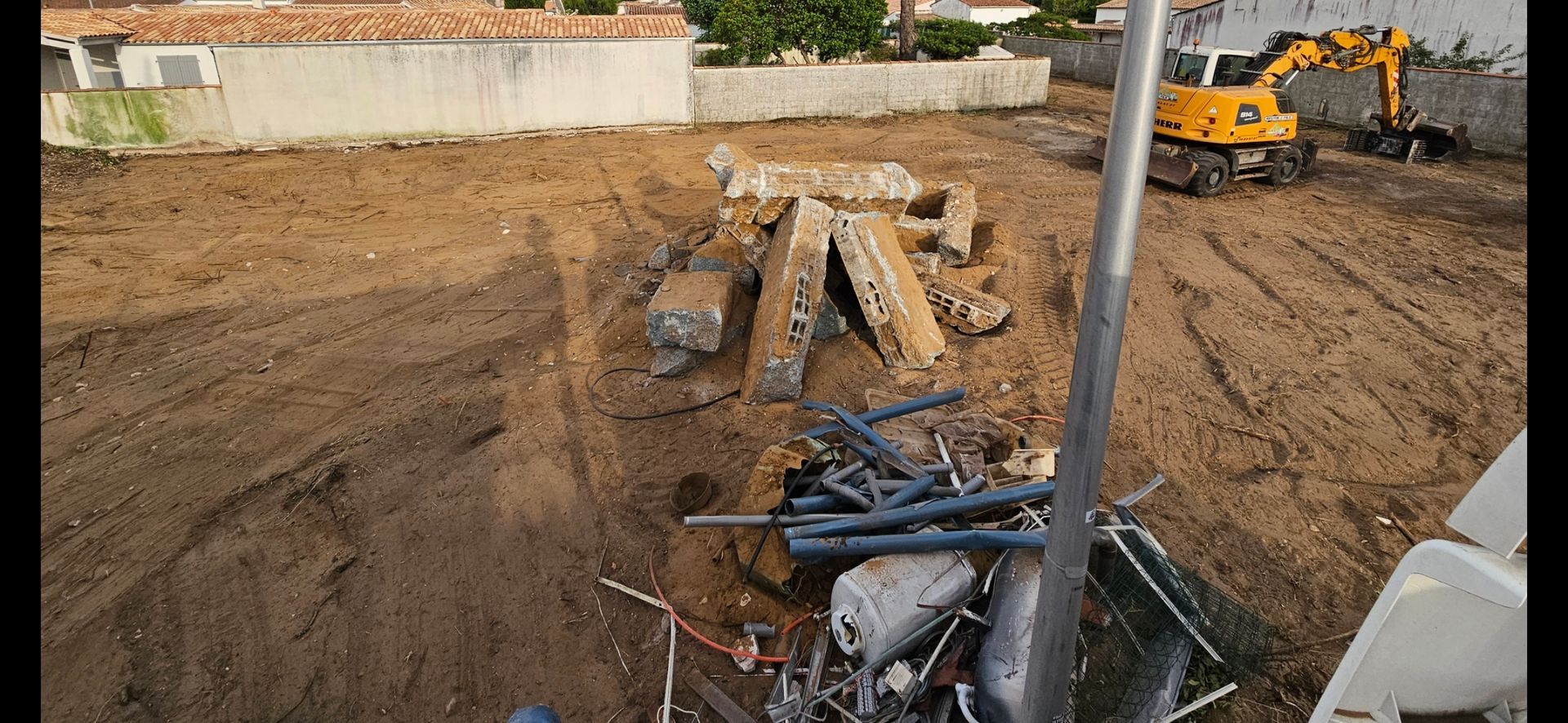 Chantier de construction avec une excavatrice, des débris et de la terre. Des bâtiments se dressent à l'arrière-plan.