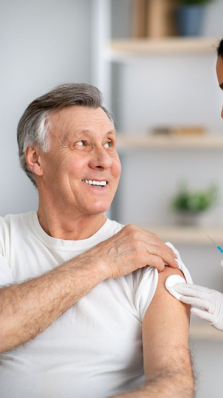 Un homme âgé vêtu d'un t-shirt blanc reçoit une injection de vaccin dans le bras par une infirmière portant des gants.