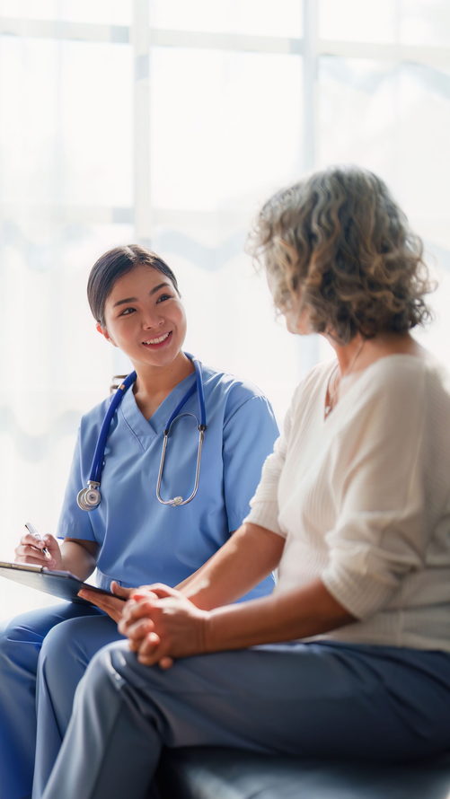Une infirmière en tenue bleue sourit à un patient âgé assis. Cadre intérieur lumineux.
