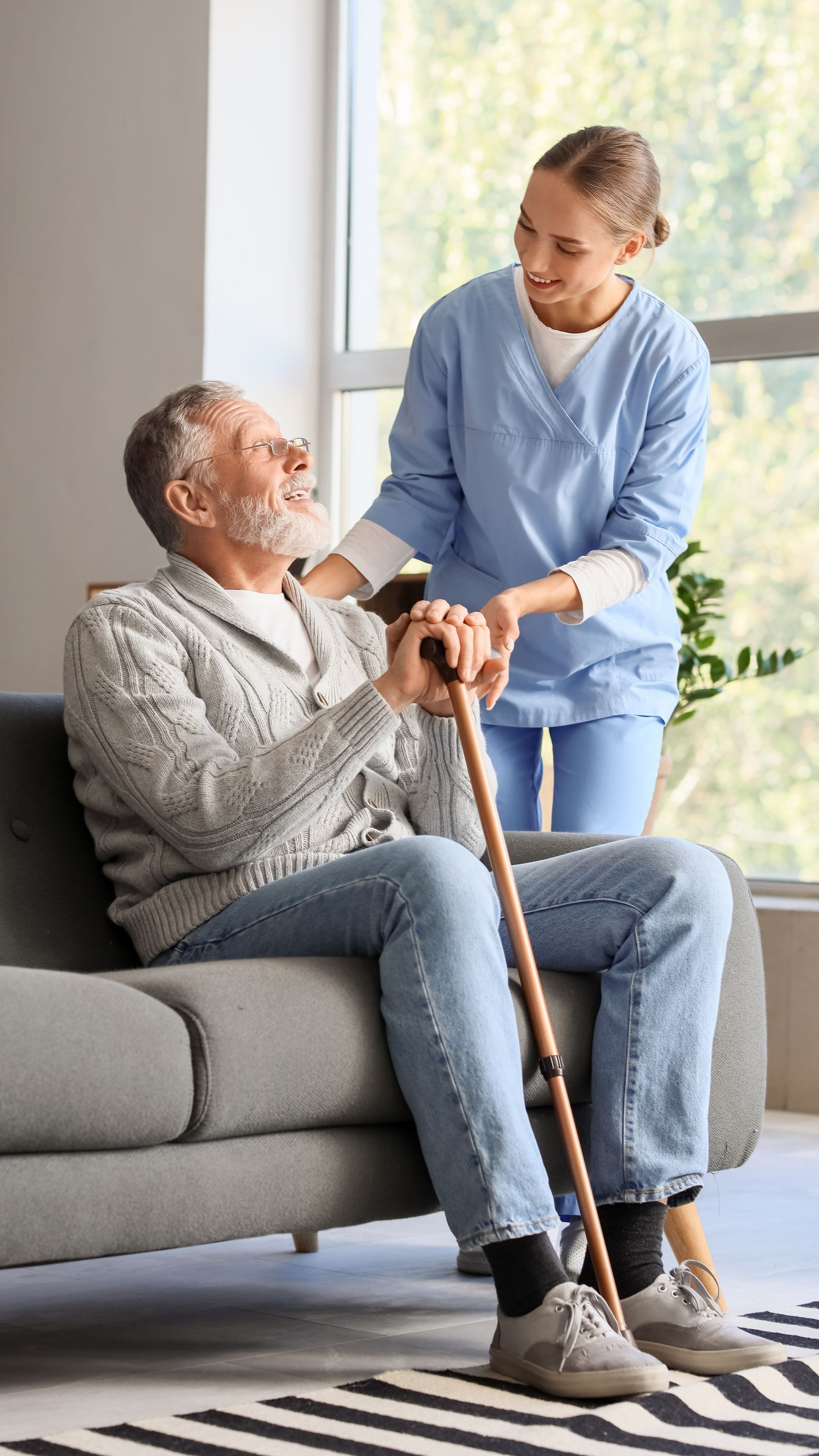 Une femme en tenue médicale bleue aide un homme âgé assis sur un canapé avec une canne. Ils sourient, à l'intérieur.