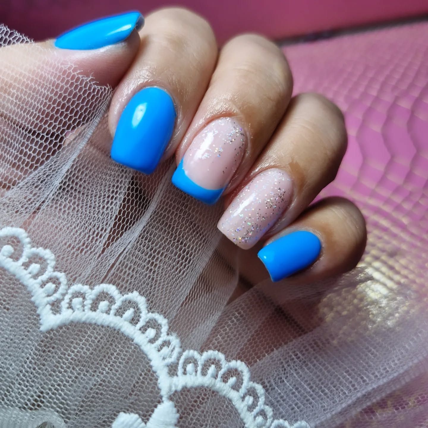 Mano con uñas azul brillante y nude con purpurina, apoyada sobre tela rosa y encaje blanco