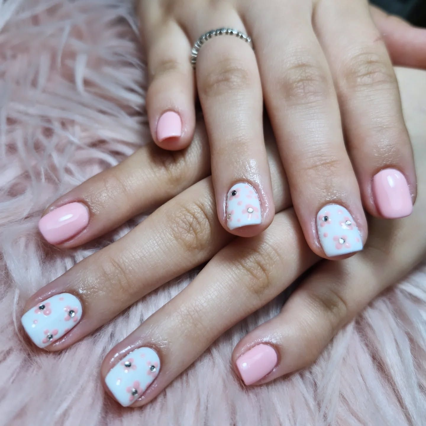 Manos con un diseño floral en tonos pastel rosa y blanco sobre las uñas, apoyadas en un fondo rosa esponjoso.
