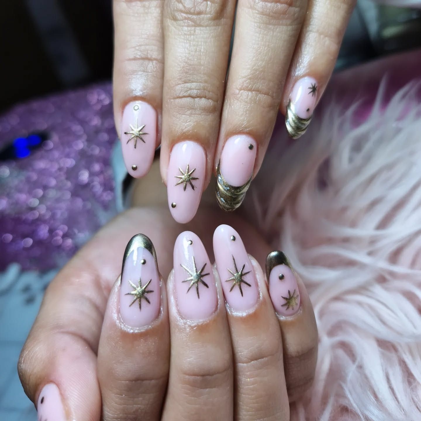Manos con uñas cuidadas de color rosa pálido y diseños de estrellas doradas, sobre un fondo morado mullido