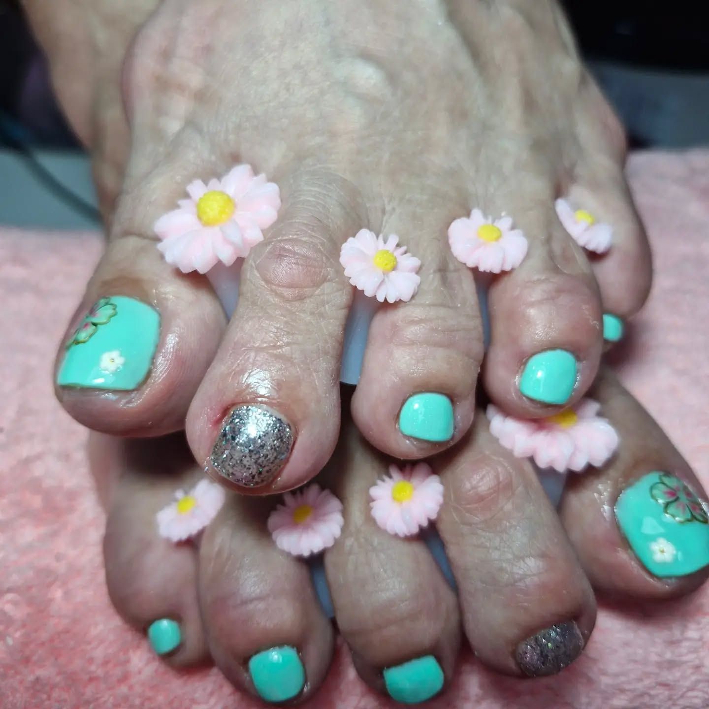 Manos con esmalte de uñas turquesa y plateado con purpurina, decoradas con pequeñas margaritas rosas