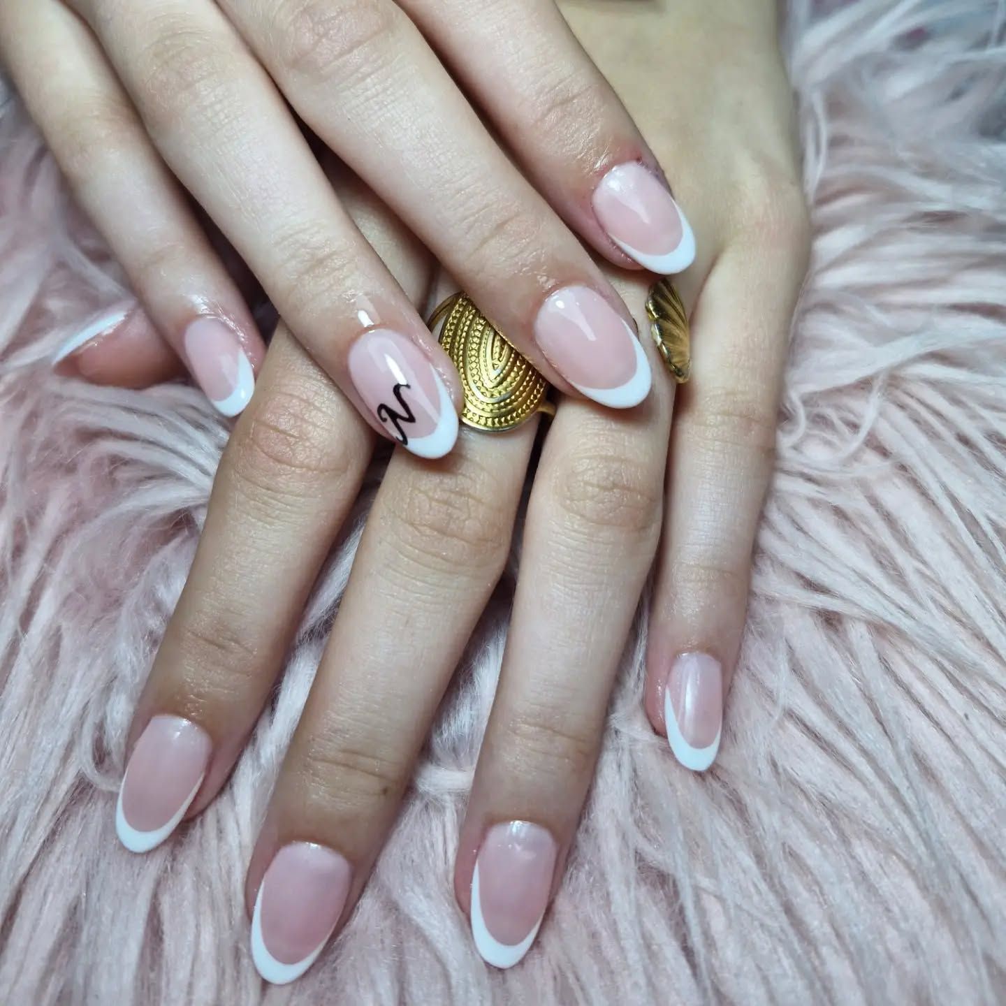 Unas manos con uñas francesas de punta rosa pálido sostienen un anillo dorado con textura sobre un fondo de pelo rosa suave