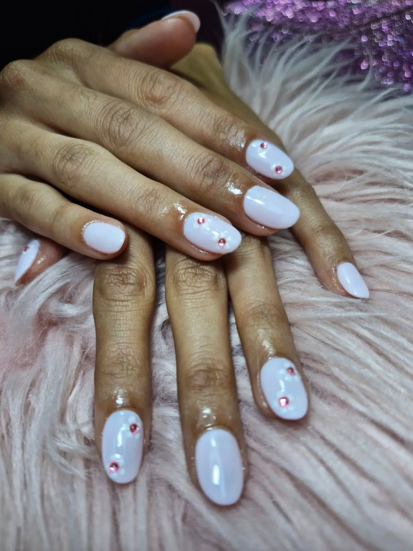 Manos con uñas cuidadas de color rosa pálido y pequeños detalles de strass, apoyadas sobre una superficie morada y mullida
