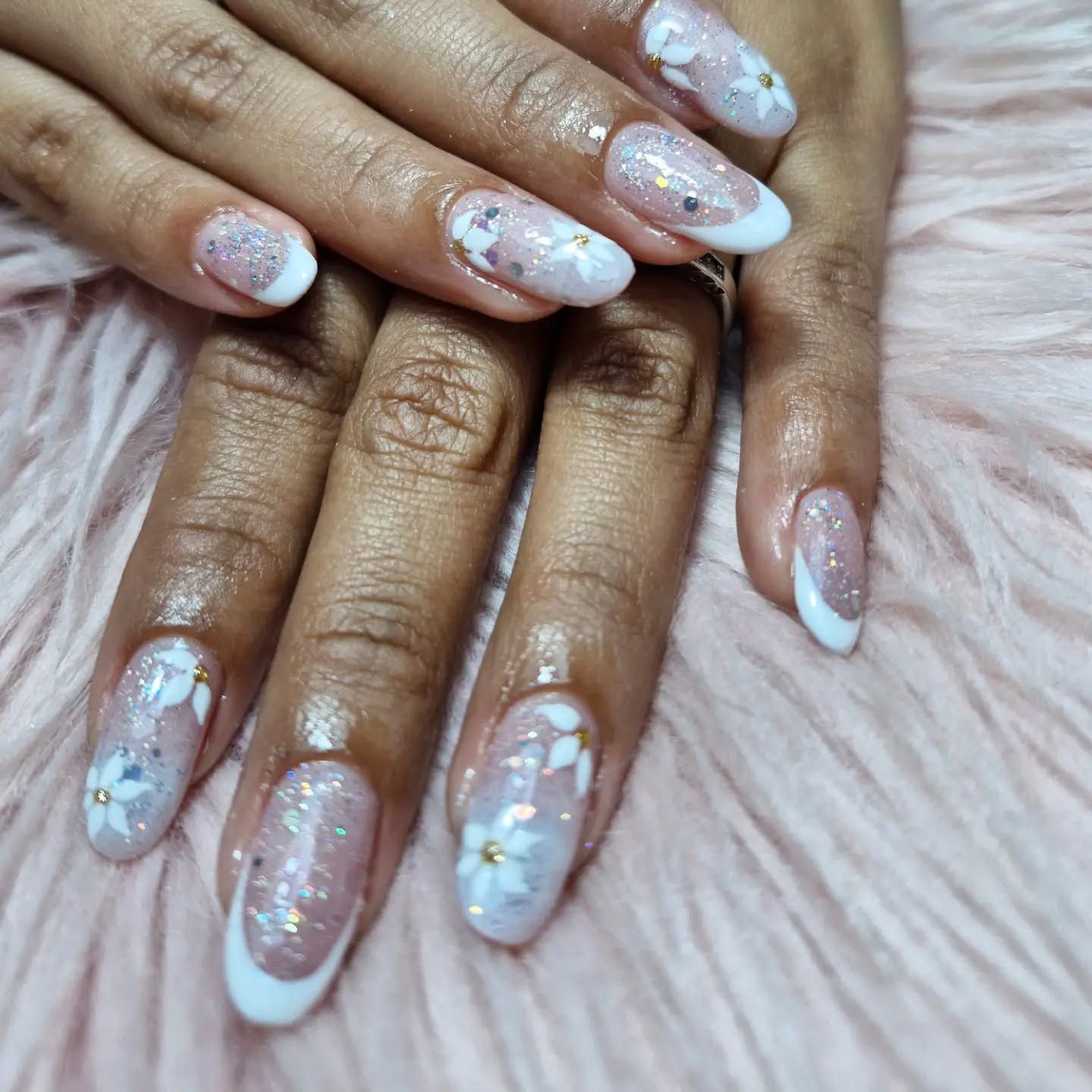 Manos con diseños florales iridiscentes y blancos en las uñas, sobre un fondo rosa suave con textura