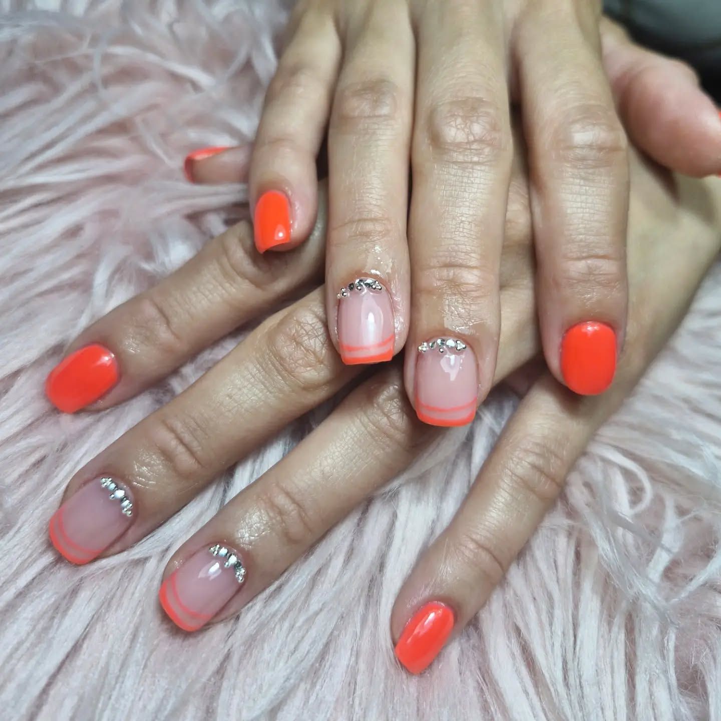 Manos con uñas en degradado coral anaranjado y nude, con detalles de pedrería plateada, sobre un fondo rosa y esponjoso