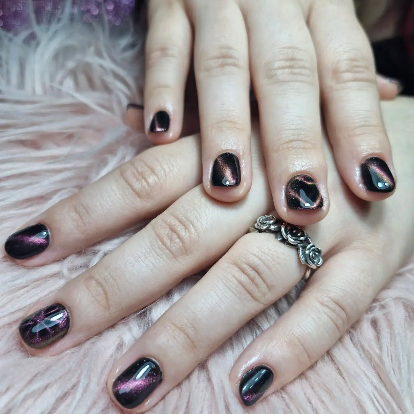 Manos con esmalte de uñas negro iridiscente sobre pelo sintético rosa, con un anillo plateado.