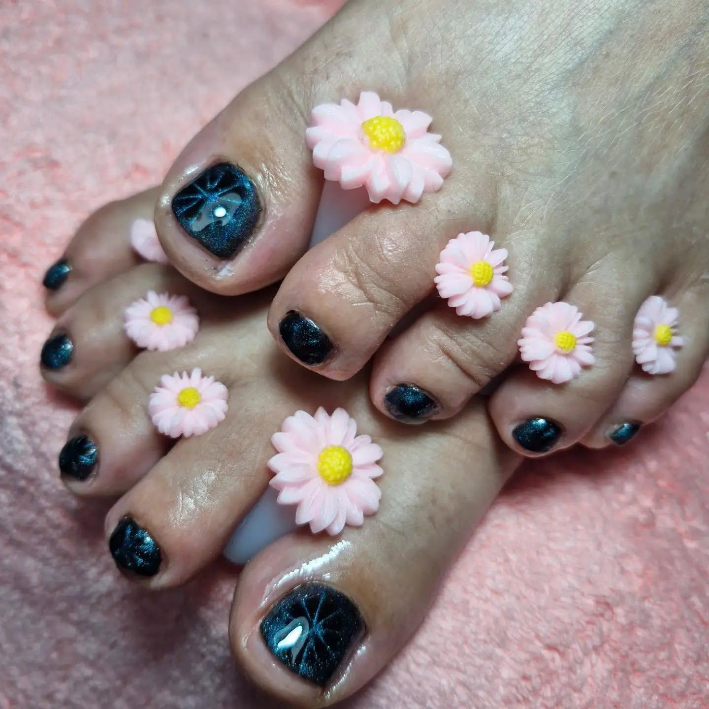 Dedos de los pies con pedicura, esmalte negro con purpurina y margaritas rosas en las uñas, sobre un fondo rosa