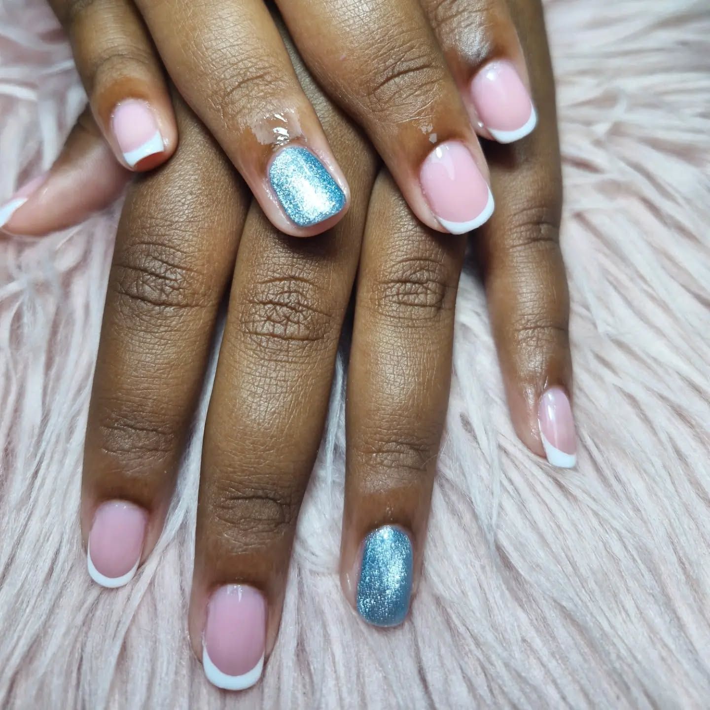 Manos con uñas de color rosa pastel y dos uñas decorativas con purpurina azul sobre un fondo blanco suave