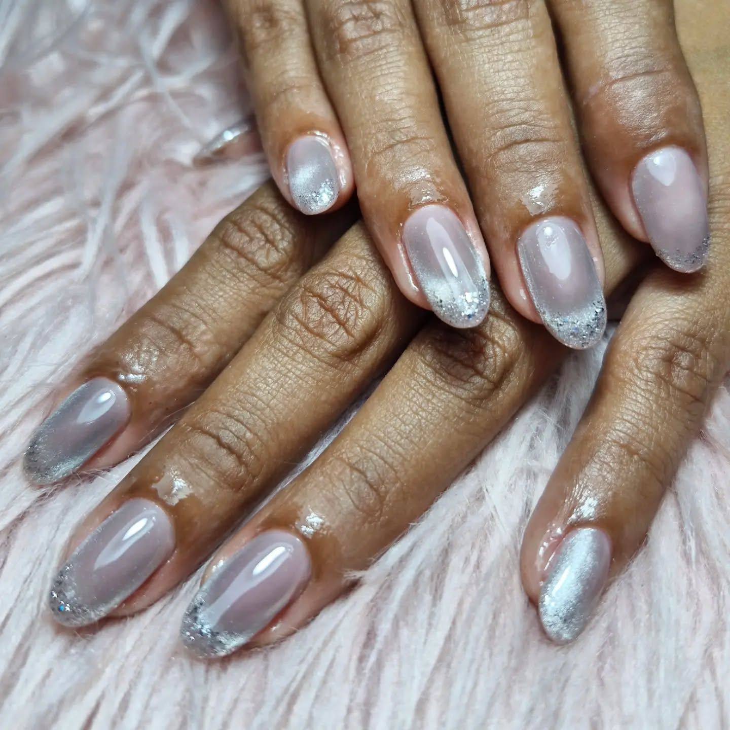 Manos con uñas cuidadas en tonos lavanda brillante y plateado sobre un suave fondo rosa y peludo