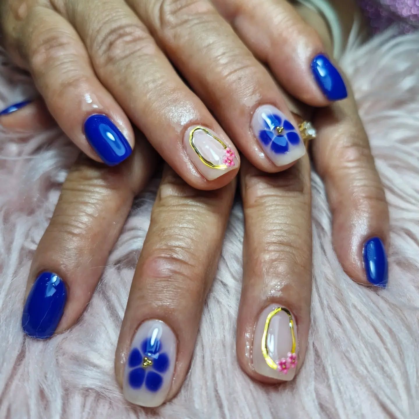 Manos con uñas decoradas con motivos florales en azul real sobre un fondo rosa suave y peludo
