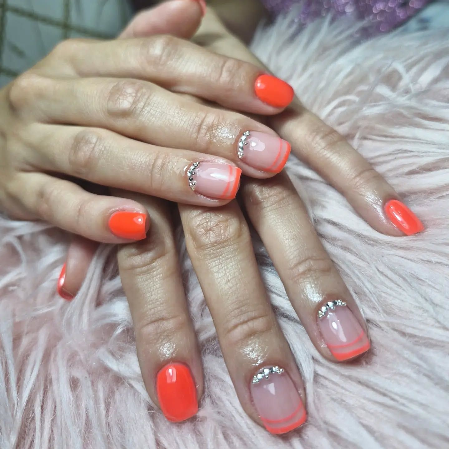 Manos con las uñas arregladas con purpurina coral y rosa, apoyadas sobre una tela mullida.