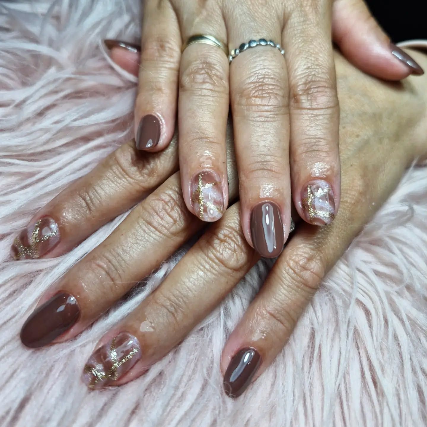 Manos con uñas marrones brillantes y decoradas con purpurina, apoyadas sobre piel sintética rosa.