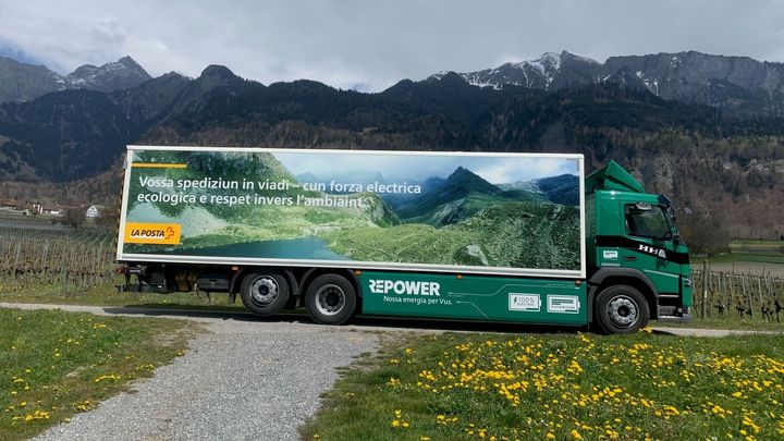 Ein grüner Lastwagen fährt auf einer unbefestigten Straße durch ein Feld mit Bergen im Hintergrund.