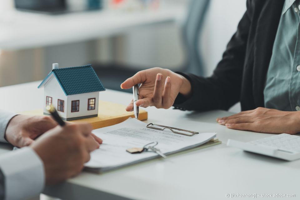 Eine Person unterzeichnet ein Dokument, eine andere zeigt mit einem Stift darauf; auf dem Tisch stehen ein Modellhaus, Schlüssel und Gläser.