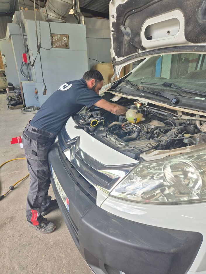 Un mécanicien en chemise noire et pantalon de travail gris travaille sur le moteur d'une camionnette blanche dans un garage.
