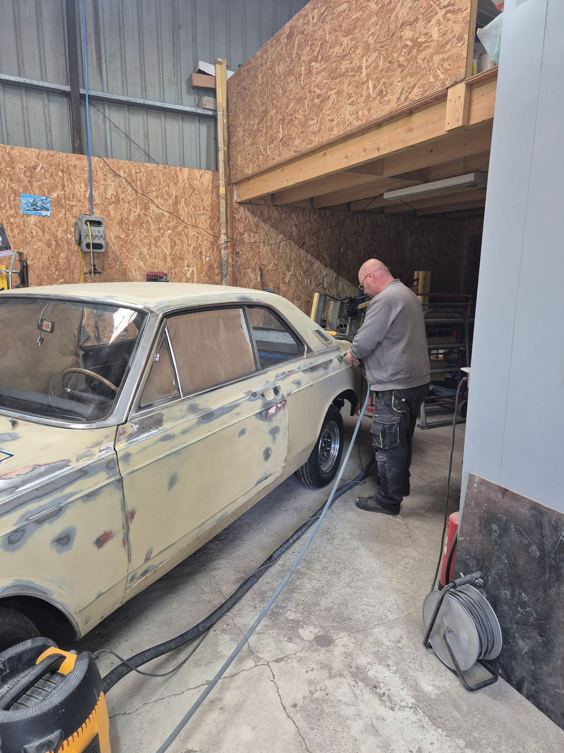 Dans un atelier aux murs lambrissés et dotés d'une mezzanine, une personne ponce, une voiture ancienne jaune pâle.