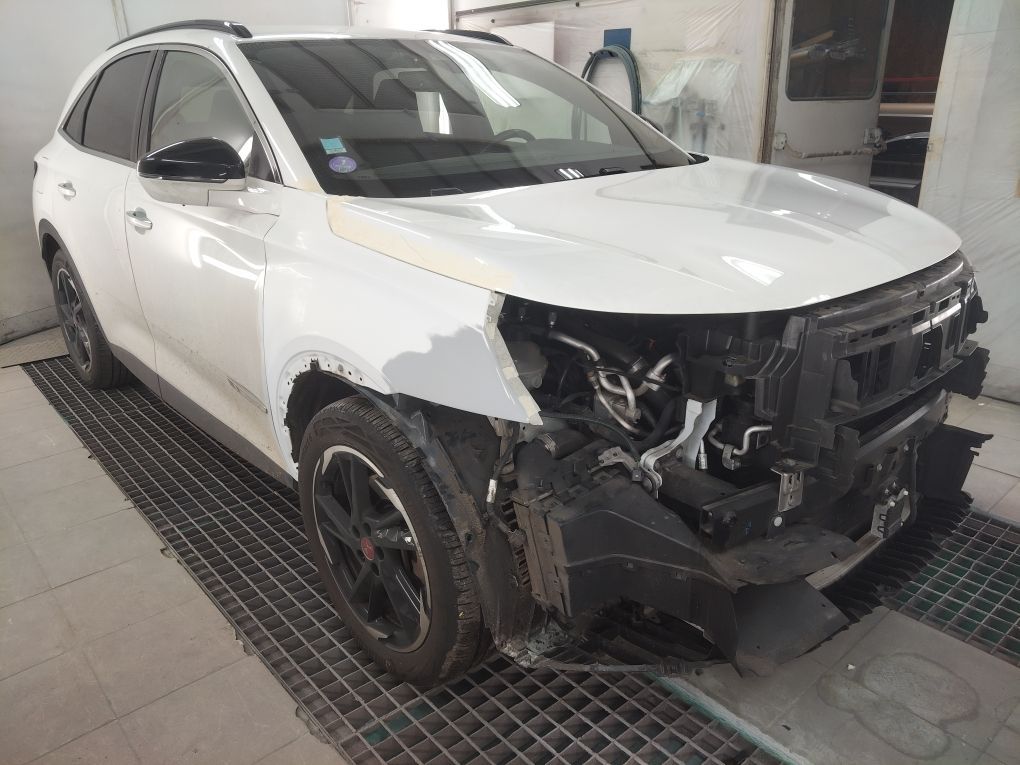 Un SUV blanc dans un atelier de carrosserie, le pare-chocs avant et le phare démontés, laissant apparaître du mastic gris sur l'aile.