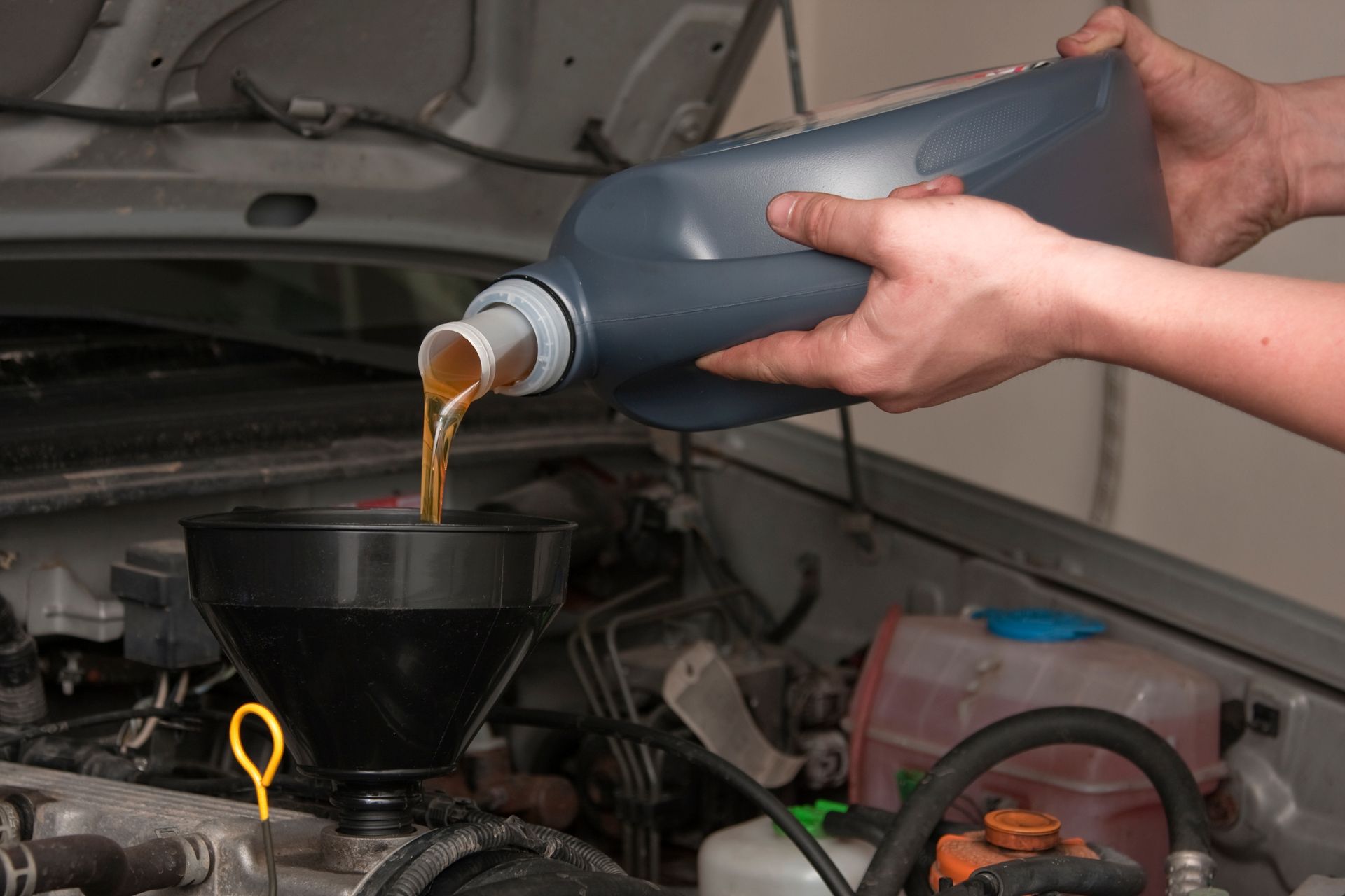 Une personne verse de l'huile à moteur dorée d'une bouteille en plastique grise à l'aide d'un entonnoir noir dans un moteur de voiture.