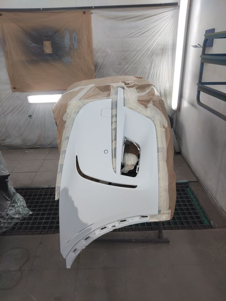 Un pare-chocs de voiture blanc, masqué avec du papier et du ruban adhésif, est monté sur un support dans un atelier de peinture automobile professionnel.