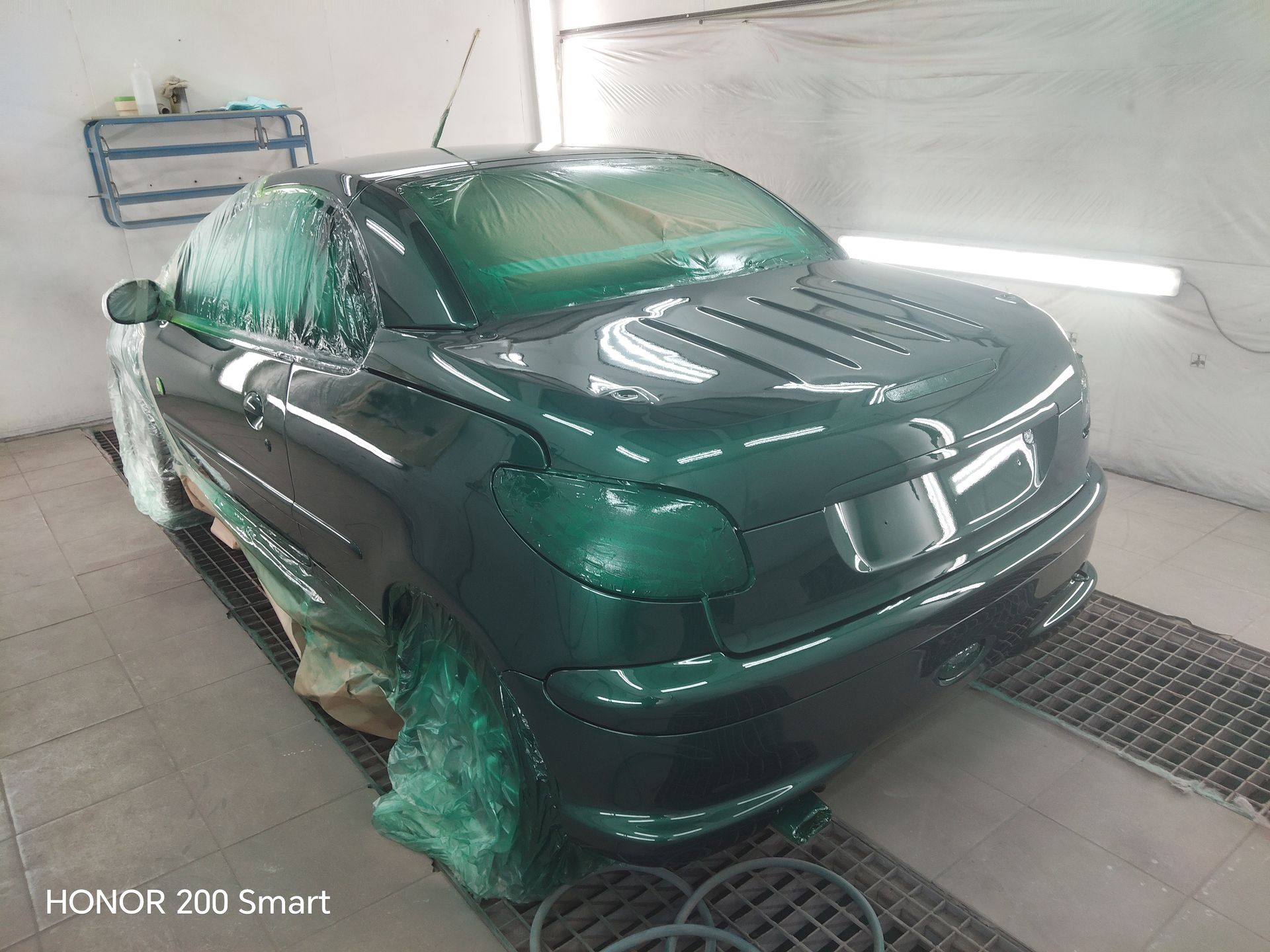 Une voiture vert foncé à l'intérieur d'une cabine de peinture, ses vitres étant recouvertes de papier protecteur pour une nouvelle application de peinture.