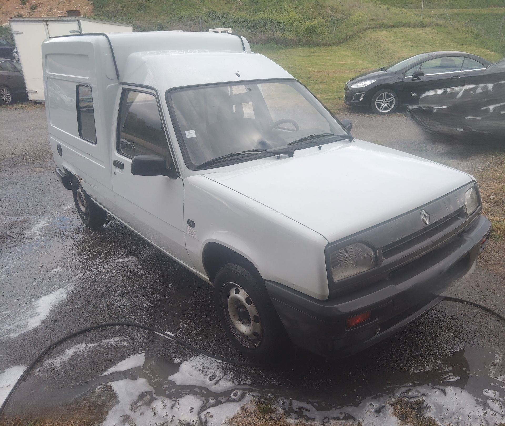Fourgonnette blanche garée sur un terrain gravillonné et humide, avec une autre voiture en arrière-plan.