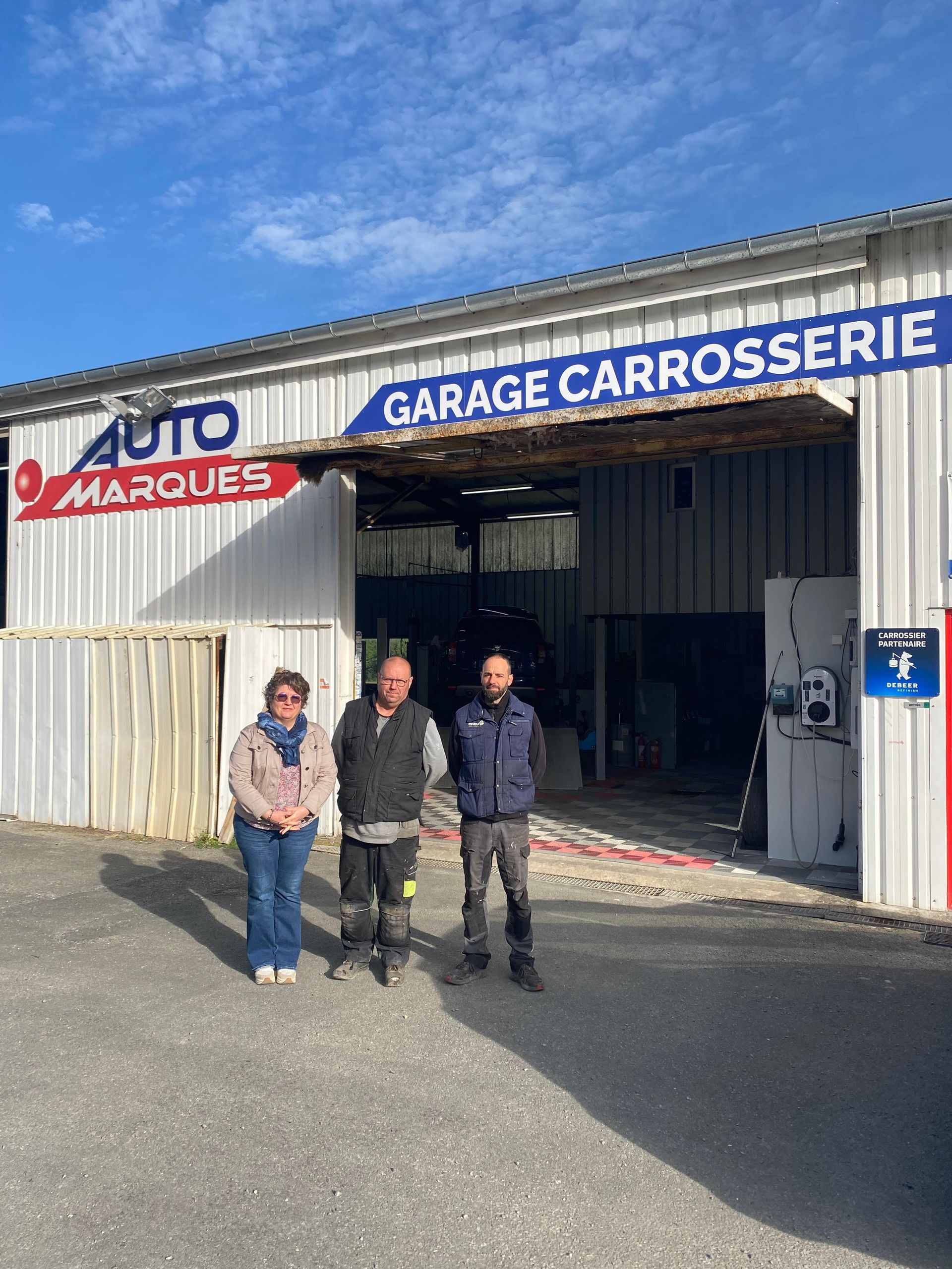 Trois personnes se tiennent devant un garage de carrosserie avec une enseigne bleue par une journée ensoleillée