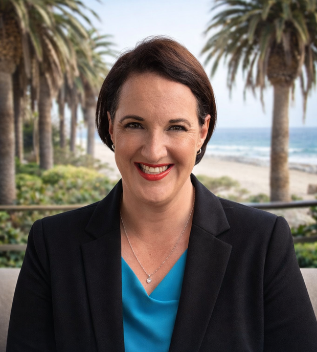 Woman in blazer smiles at the camera, blue top, beach background.