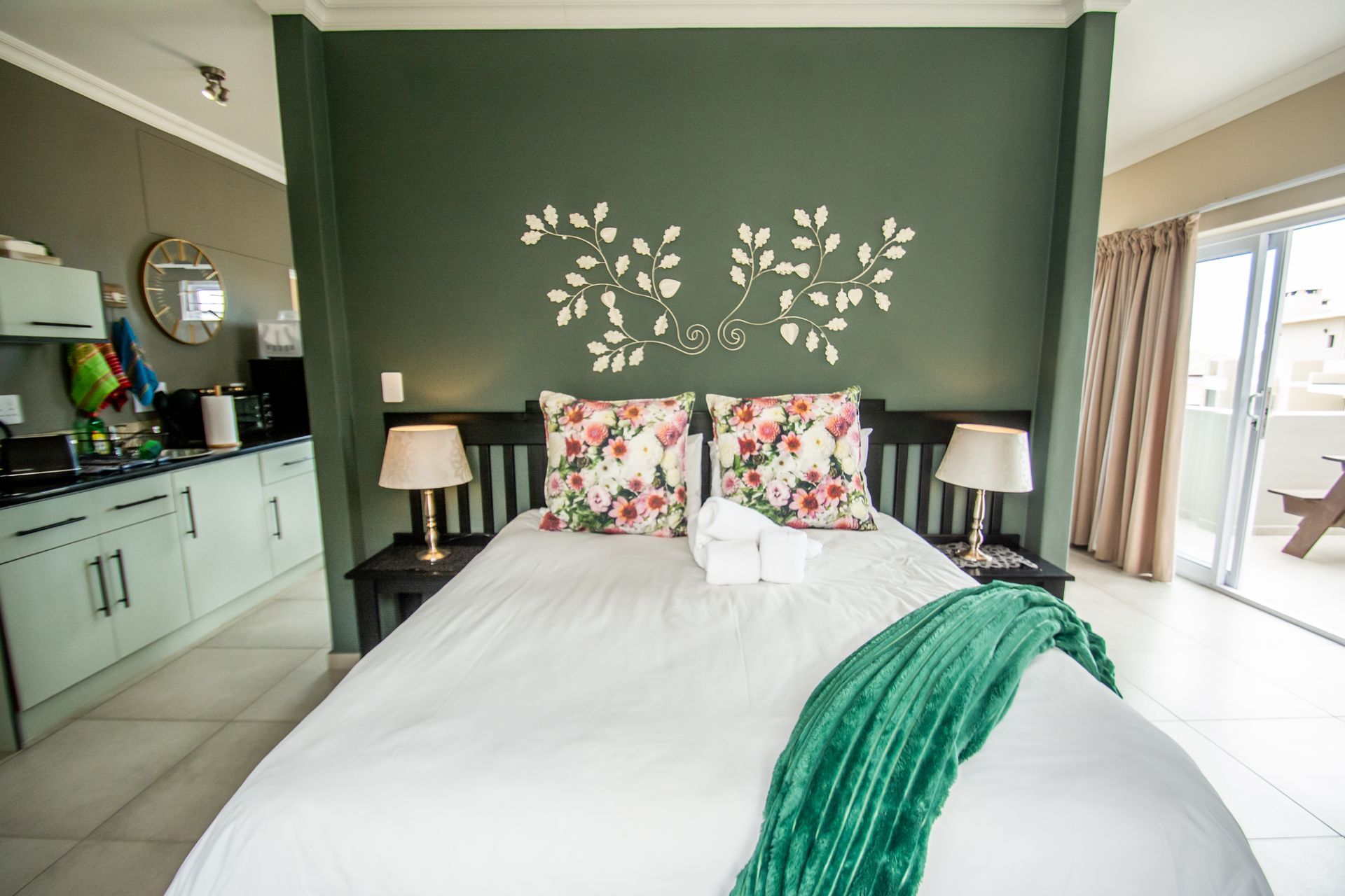 Bedroom with a queen bed, green accent wall, small kitchen, and a balcony.