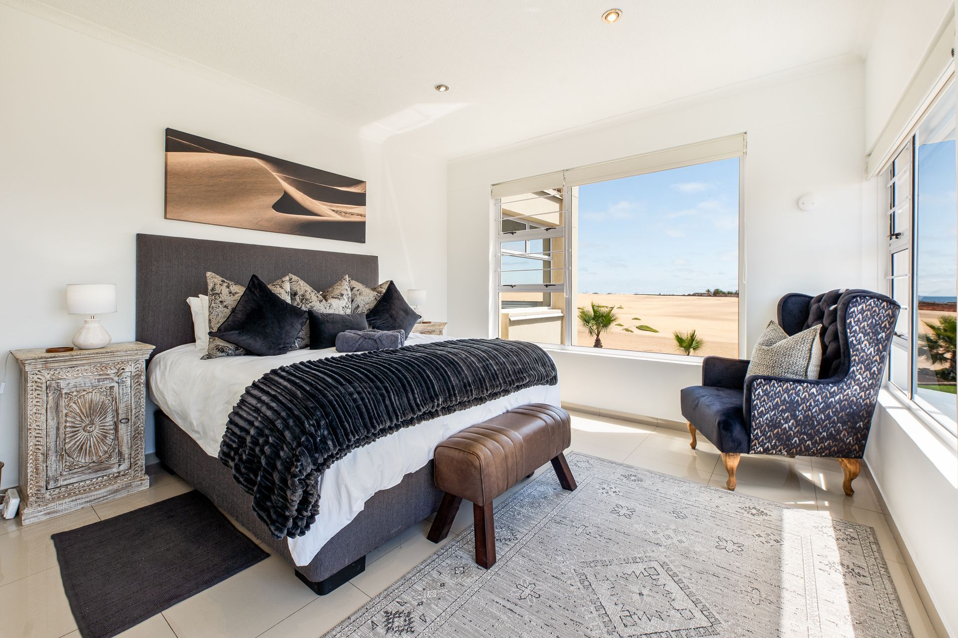 Bedroom with bed, patterned rug, armchair, and window overlooking a desert landscape.
