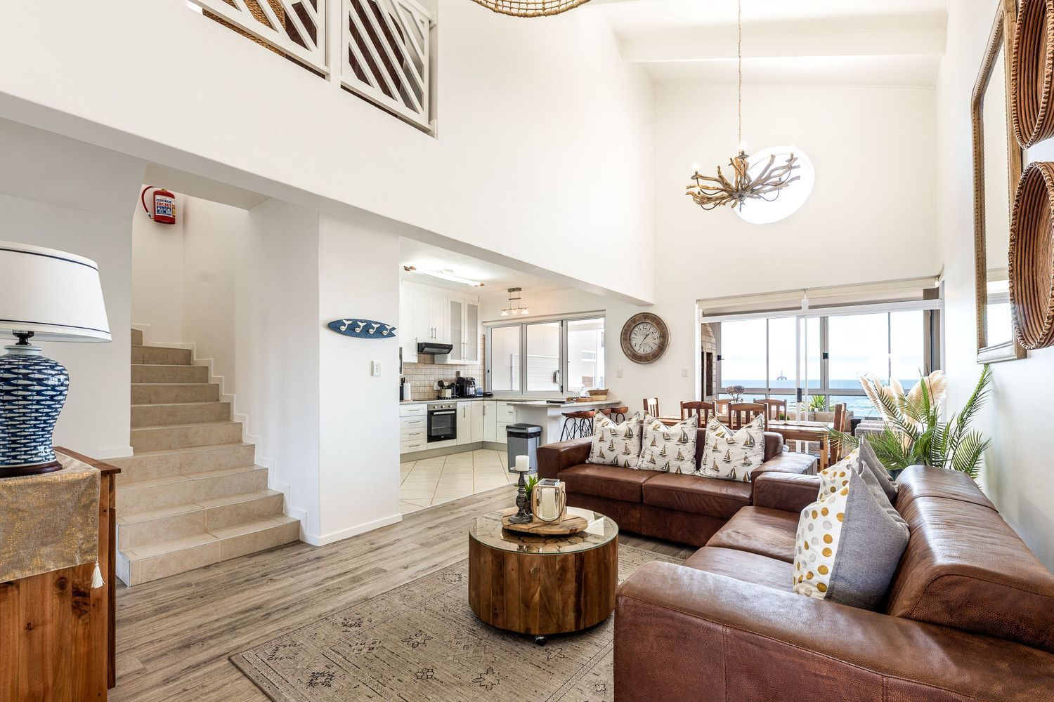 Living room with brown leather sofa, rug, stairs, and kitchen visible.