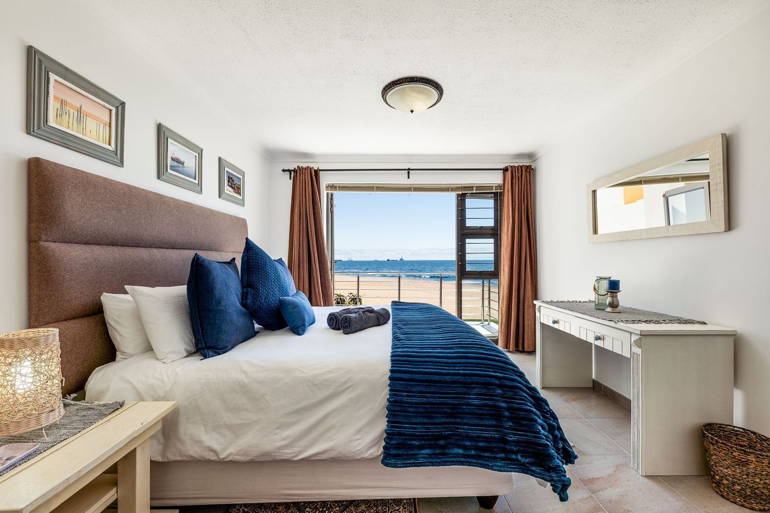 Bedroom with a bed, ocean view, and white desk.  Dark blue accents, tan headboard and curtains.