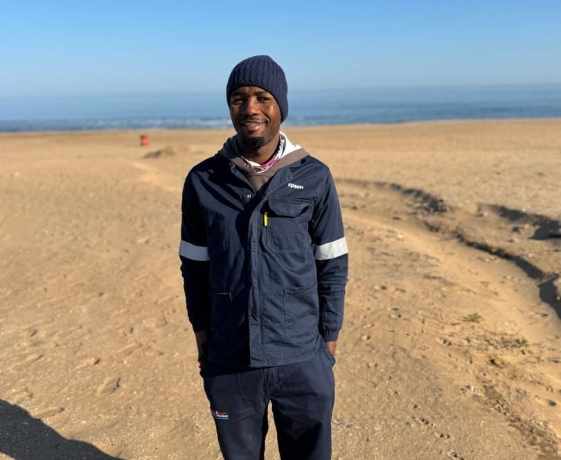 Man standing on a sandy beach, wearing a blue jacket and beanie, with the ocean in the background.