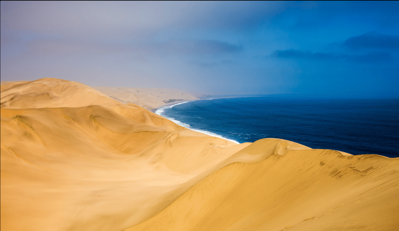 Sandy desert dunes meeting the blue ocean under a cloudy sky.