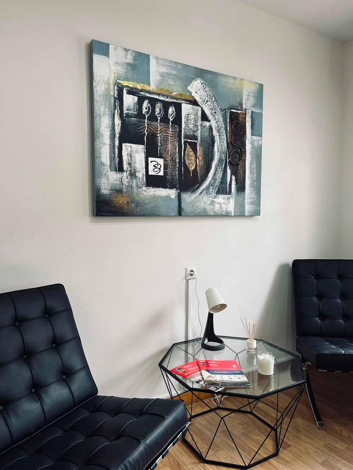 Sala de estar de oficina moderna con dos sillas negras, mesa de cristal y arte abstracto en la pared.