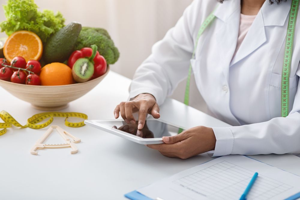 Una mujer está usando una tableta mientras está sentada en una mesa con un tazón de frutas y verduras.