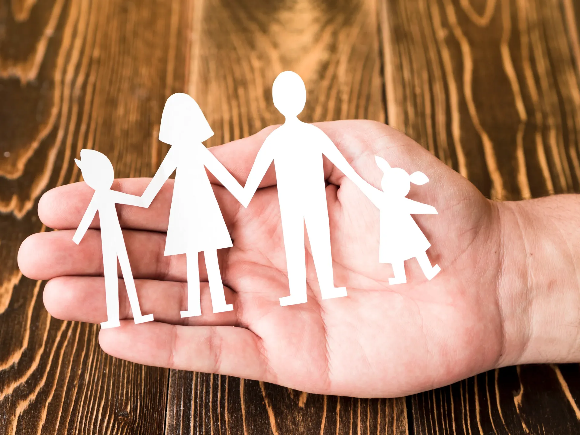 Una mano sostiene una figura de papel recortada que representa a una familia de pie en fila sobre un fondo de madera.
