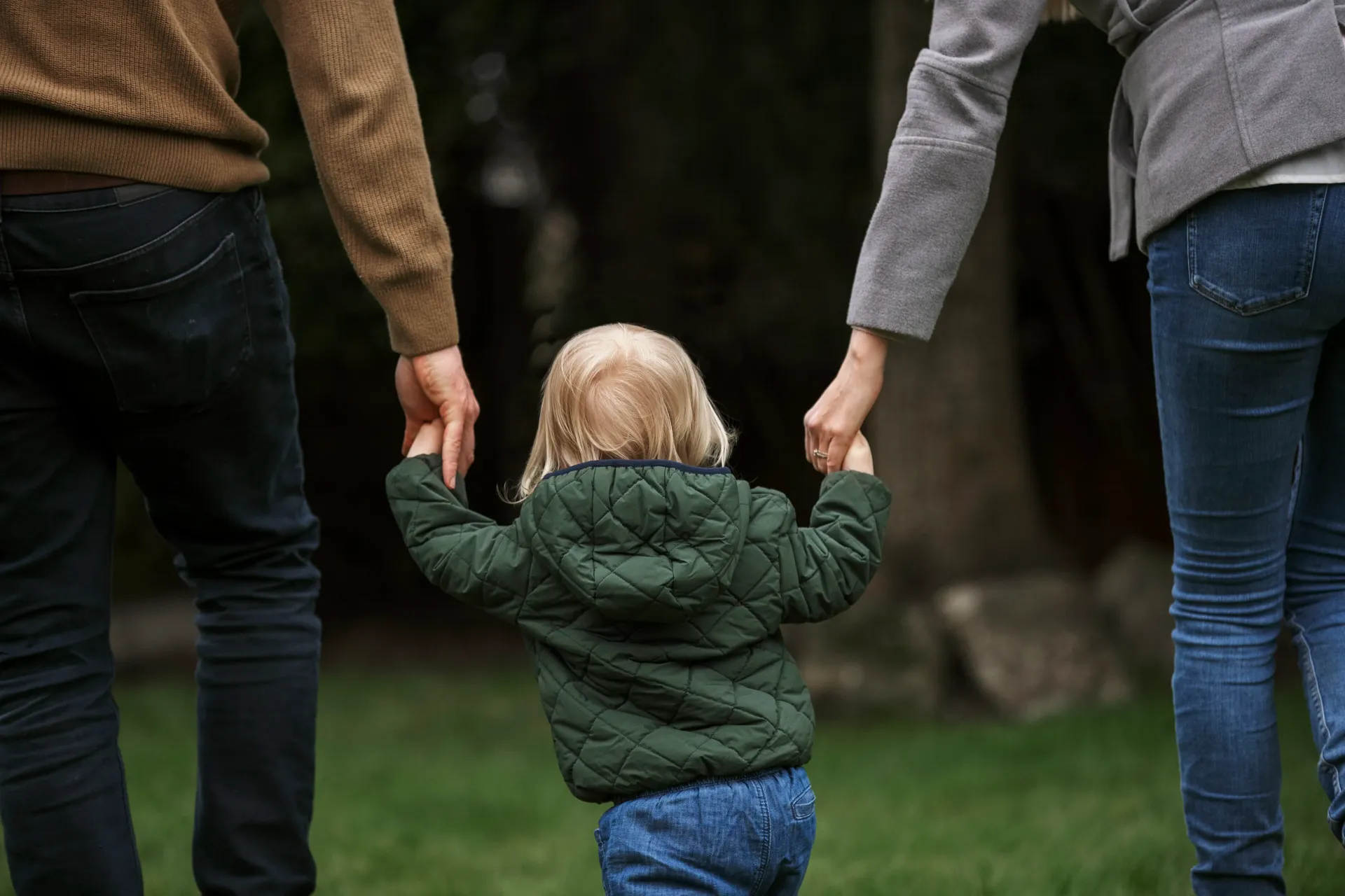 Un niño pequeño con una chaqueta acolchada verde camina al aire libre, de la mano de dos adultos, sobre un césped.