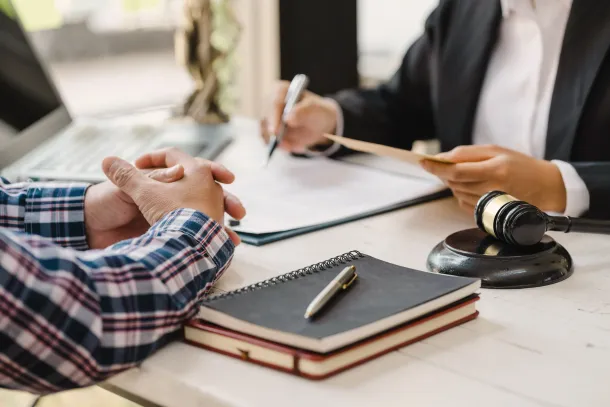 Un profesional vestido de traje revisa documentos en un escritorio frente a un cliente; un mazo de madera yace cerca.