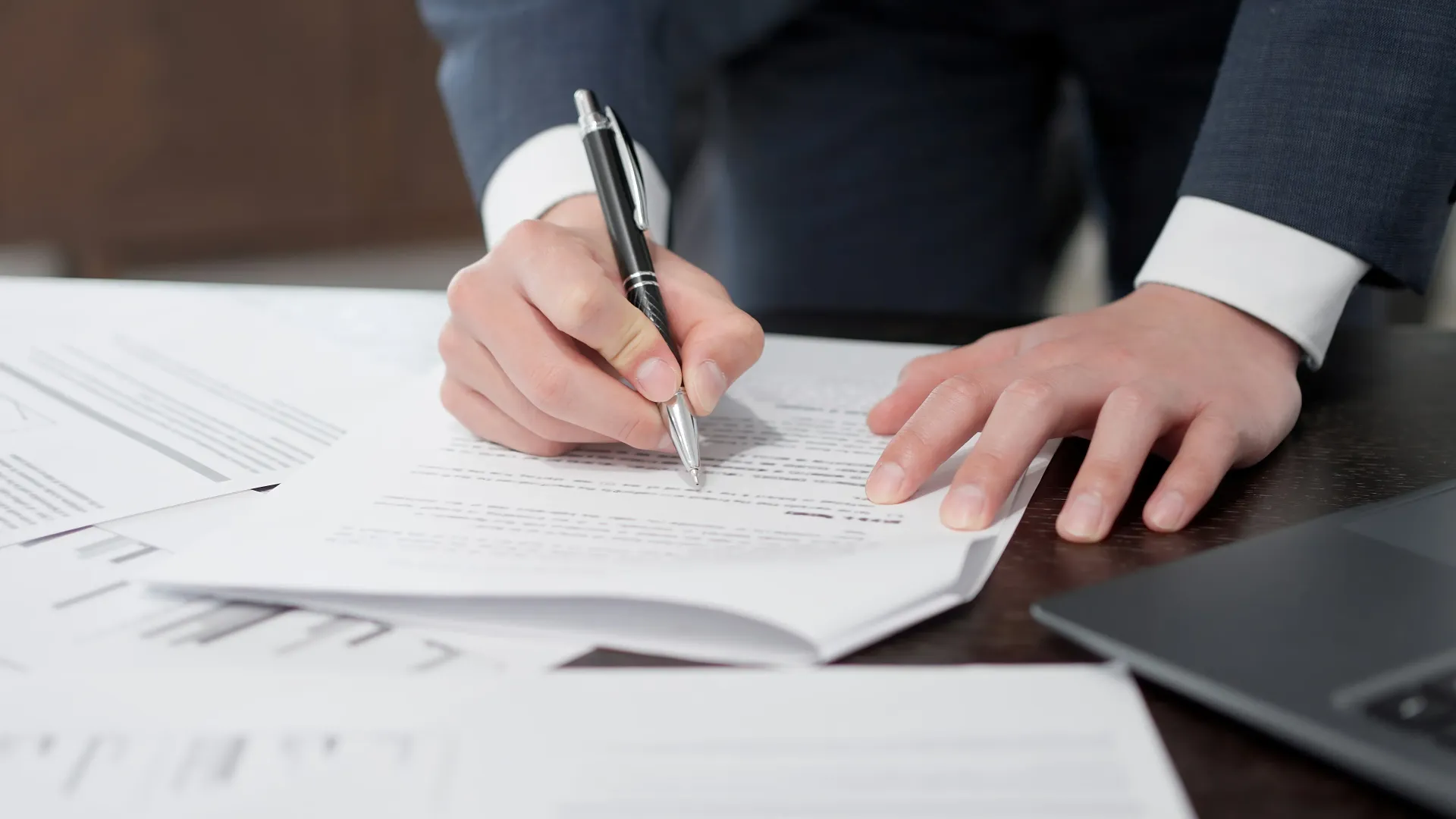 Una persona vestida con traje de negocios escribe en un documento con un bolígrafo, con papeles adicionales y una computadora portátil sobre el escritorio.