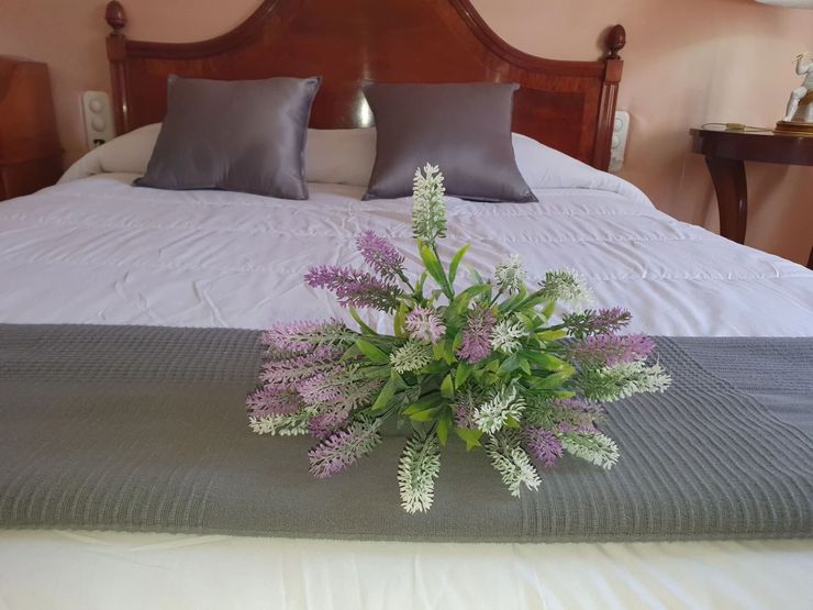 Dormitorio con colcha blanca, almohadas grises y un centro de mesa floral morado y blanco a los pies de la cama.