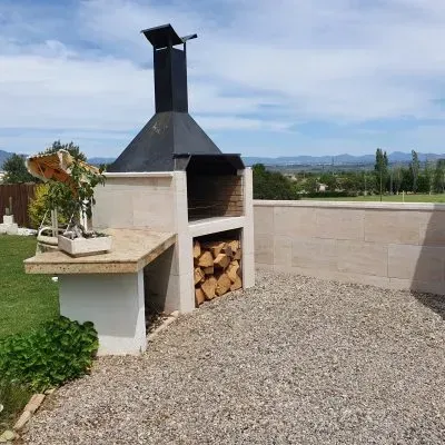 Patio exterior con parrilla de ladrillo integrada, leña apilada y suelo de grava con vistas a un paisaje lejano.