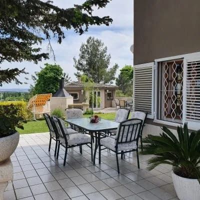 Patio con mesa y sillas de cristal junto a una casa, con vistas a un jardín y a las colinas lejanas.