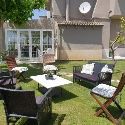 Zona de estar en el patio con sillas negras, mesa blanca y sofá en un jardín trasero cubierto de hierba junto a una casa.