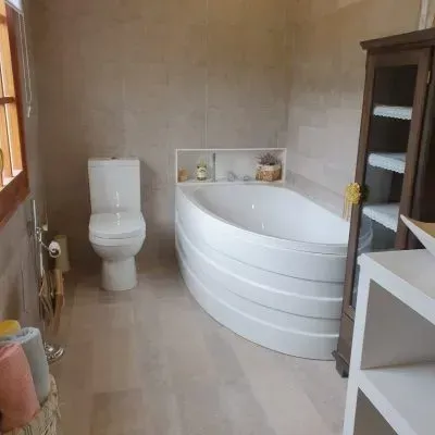 Baño moderno con bañera de esquina, inodoro, paredes alicatadas y estantería de madera.