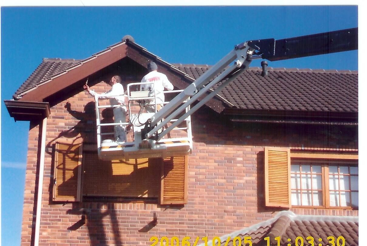 Dos trabajadores pintan el revestimiento de una casa desde una plataforma elevadora. Edificio de ladrillo rojo, molduras marrones y cielo azul.