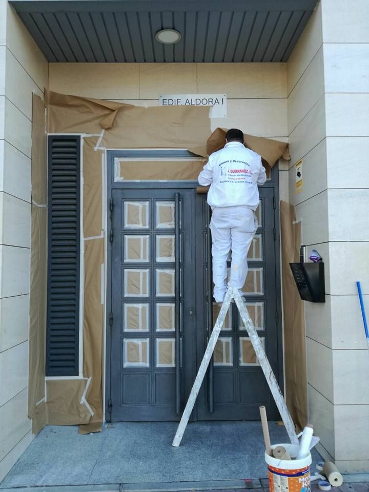 Persona en una escalera pintando una puerta gris. Papel marrón cubre las paredes circundantes. Escenario exterior.